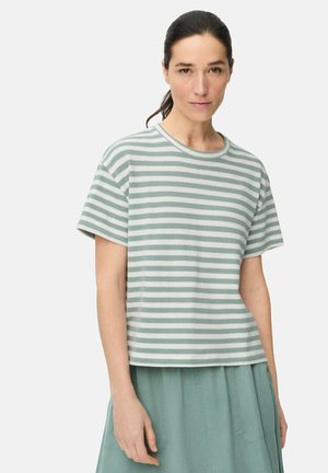 Kvinde med mørkt hår sat tilbage iført en lysegrøn og hvid stribet kortærmet skjorte og lysegrøn nederdel mod en hvid baggrund.