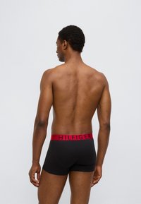 Μαύρο ανδρικό εσώρουχο τύπου boxer με κόκκινη ελαστική μέση που φέρει το λογότυπο "HILFIGER" σε μαύρο. Το υλικό φαίνεται λείο· ο σχεδιασμός είναι εφαρμοστός και μοντέρνος.