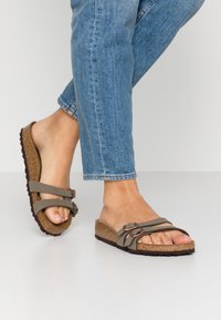 Fødder iført khaki grønne sandaler med dobbelts spænde og korksåler, sammen med blå jeans på en lys grå baggrund.