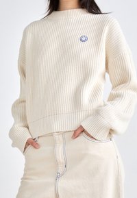 Pull crème en tricot côtelé avec un col rond et un logo sourire bleu sur la poitrine ; accompagné d'une jupe beige taille haute avec des surpiqûres bleues.