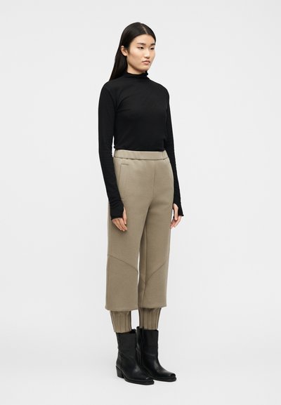 Γυναίκα με μακριά σκούρα μαλλιά φοράει μαύρο πουλόβερ με ζιβάγκο, μπεζ παντελόνι cropped με ριμπ μανσέτες και μαύρα μποτάκια μέχρι τον αστράγαλο, στέκεται σε λευκό φόντο.