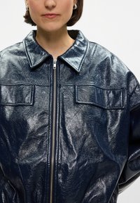 Jūras zila mākslīgās ādas jaka ar rāvējslēdzēju, lielu apkakli un divām priekšējām kabatām. Iezīmējas ar spīdīgu tekstūru un nemanāmām krokām.