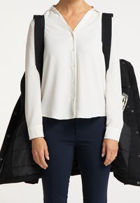 Blouse blanche à manches longues avec col et fermeture à boutons. Portée avec un pantalon marine, accompagnée d'un manteau matelassé noir posé sur une épaule.