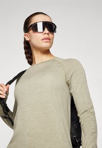 Atletska odjevna majica dugih rukava u maslinasto zelenoj teksturiranoj tkanini. Model nosi tamne oversized sunčane naočale i drži crnu torbu na ramenu.