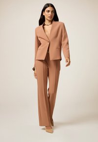 Costume beige ajusté avec un blazer à encolure en V profonde et un pantalon à jambes larges. Tissu lisse, bouton doré unique et poches discrètes. Des talons beiges complètent le look.