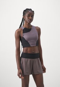 Sportoutfit mit einem figurbetonten lila und schwarzen Cropped-Top, kombiniert mit lockeren braunen Shorts. Der Stoff wirkt glatt mit kontrastierenden Einsätzen.