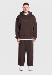 Brązowa bluza z kapturem z kieszenią z przodu, z haftem "Prosto." w zestawie z dopasowanymi brązowymi spodniami dresowymi i białymi sneakerami. Gładkie tło.