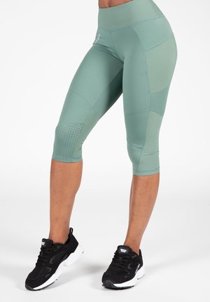 Hoge taille, mintgroene leggings hebben een gladde textuur met netaccenten en perforaties in de buurt van de kuiten. Gecombineerd met zwarte sportschoenen.