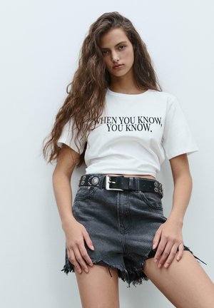 Weißes Baumwoll-T-Shirt mit schwarzem Text "WENN DU ES WEISST, WEISST DU ES." kombiniert mit zerrissenen schwarzen Denim-Shorts und einem genagelten schwarzen Gürtel.
