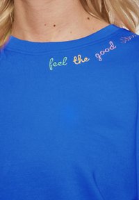 Blauwe katoenen t-shirt met een ronde hals. De halslijn heeft kleurrijke geborduurde tekst: "voel de goede vibes." Gladde textuur.