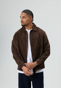 Bruine corduroy jas met een kraag, knopen aan de voorkant en lange mouwen. Gedragen over een wit shirt met een getextureerd geribbeld ontwerp.