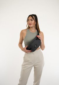 Borsa vita nera realizzata in tessuto con un design rettangolare elegante. Presenta una tasca frontale con zip e una toppa con logo. Indossata comodamente.