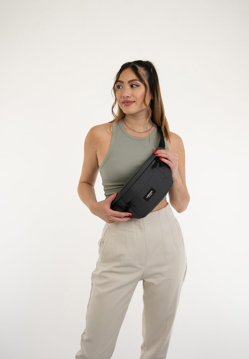 Borsa vita nera realizzata in tessuto con un design rettangolare elegante. Presenta una tasca frontale con zip e una toppa con logo. Indossata comodamente.