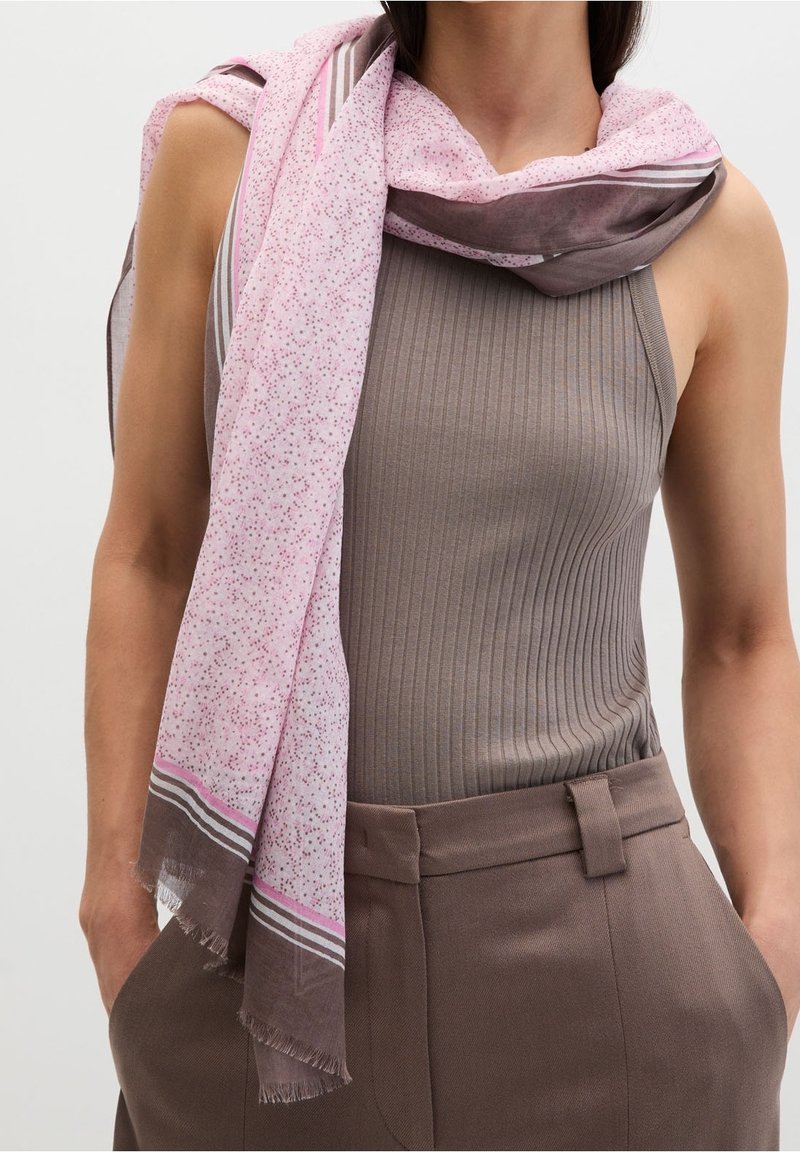 Femme portant un haut taupe côtelé sans manches et un pantalon marron cintré, avec une écharpe rose et marron à motifs drapée sur son épaule.