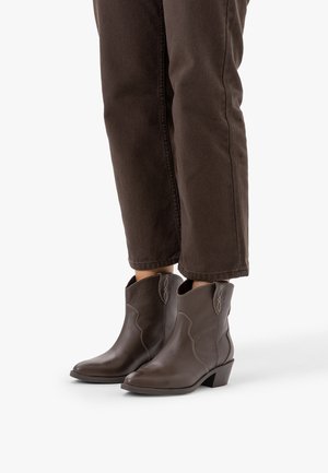 Personne portant des bottes de cowboy en cuir marron montant à la cheville avec des talons bas et un pantalon marron foncé coupé au-dessus de la cheville.