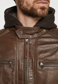 Bruine leren jas met een ritssluiting aan de voorkant, twee ritszakken op de borst en een hoodie zichtbaar eronder. Glad van textuur.