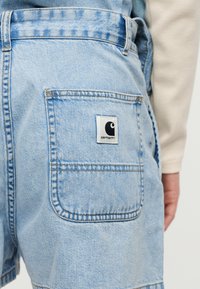 Ανοιχτό μπλε denim σορτς με τετράγωνη πίσω τσέπη που φέρει πατς με το λογότυπο Carhartt, φορεμένα από ένα άτομο με μπεζ μακρυμάνικο τοπ.