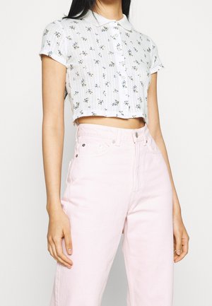 Vrouw draagt witte crop top met korte mouwen en kleine blauwe bloemenprint en hooggetailleerde pastelroze broek, staand met ontspannen armen.