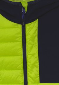 Chaqueta acolchada con cremallera frontal, presenta un brillante frontal amarillo y laterales azul marino oscuro, con una textura suave y materiales en contraste. Detalle de cremallera azul.