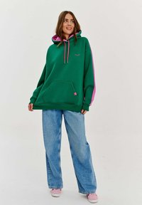 Pre veľký zelený mikinu s ružovými akcentmi, sťahovacími šnúrkami a prednou kapsou; nosená s voľnými modrými džínsami a ružovými teniskami.