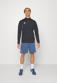 Maglia nera a maniche lunghe con zip e dettagli grigi, abbinata a pantaloni sportivi blu. Il modello indossa scarpe blu con calzini bianchi.