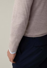 Maglione in maglia beige chiaro con toppe sui gomiti, polsini a coste con dettagli azzurri e una silhouette aderente. I pantaloni blu navy sono parzialmente visibili.