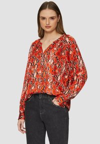 Rode blouse met slangenprint, een V-hals en lange mouwen. De stof heeft een zachte textuur en een losse pasvorm; gecombineerd met zwarte jeans met studaccenten.