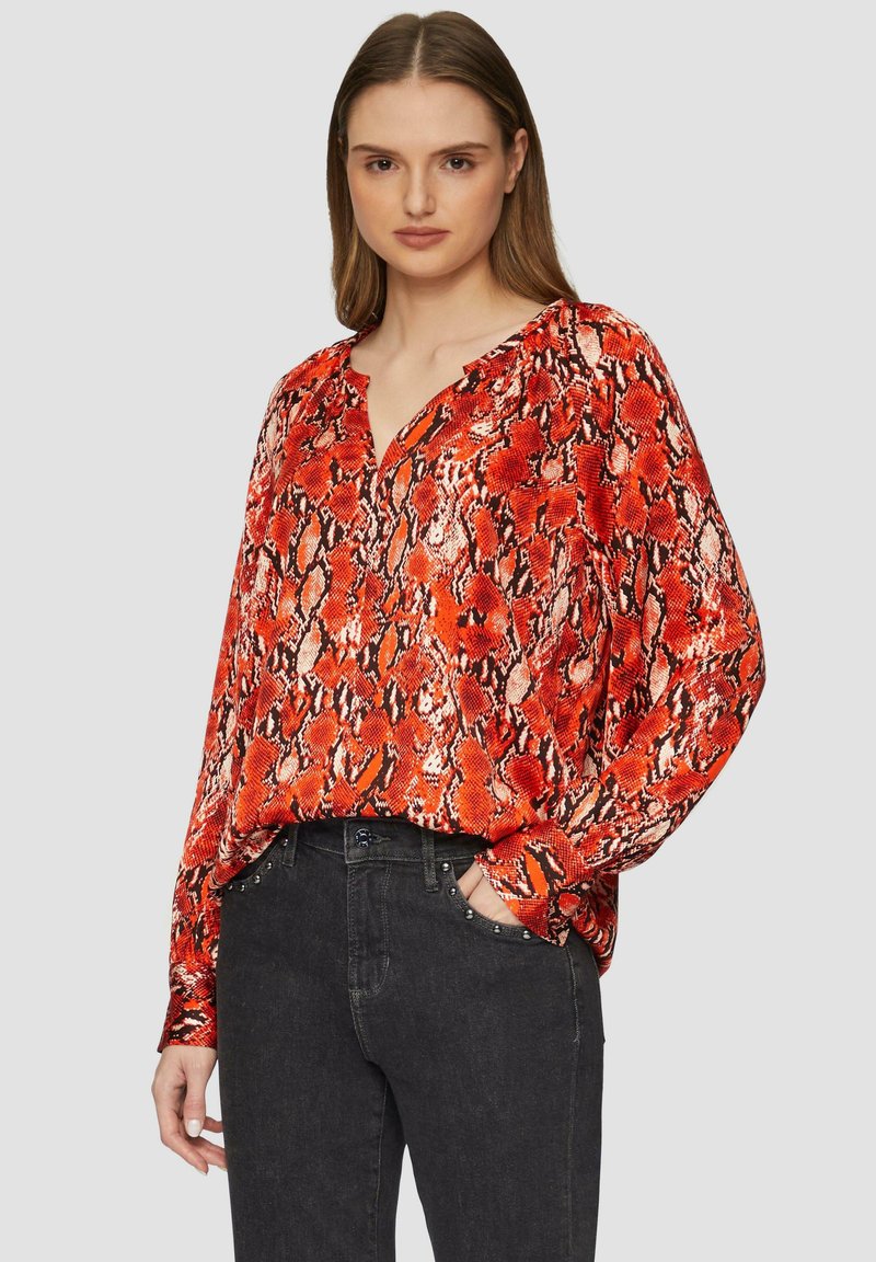 Rode blouse met slangenprint, een V-hals en lange mouwen. De stof heeft een zachte textuur en een losse pasvorm; gecombineerd met zwarte jeans met studaccenten.