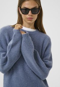 Blauer, flauschiger Strickpullover mit langen, lockeren Ärmeln und rundem Halsausschnitt. Das Modell trägt außerdem eckige schwarze Sonnenbrillen.