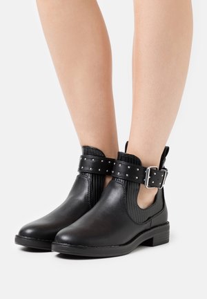 Black ankle boots with studded buckle straps, textured elastic sides, and low block heels worn on bare legs against a plain light background.