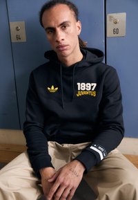 Svart hoodie med gul text "1897 JUVENTUS" och logga. Har en känguruficka och ribbade ärmslut, kombinerad med beige byxor.