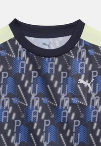 Σκούρο ναυτικό μπλουζάκι με στρογγυλή λαιμόκοψη, με μπλε και γκρι γεωμετρικά μοτίβα, επαναλαμβανόμενο κείμενο "PUMA", λευκό λογότυπο Puma και ανοιχτοκίτρινες πάνελ στους ώμους.