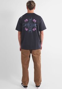 T-shirt en coton noir avec un grand graphisme de scorpions et de fleurs en bleu et rose au dos, associé à un pantalon ample de couleur sable.
