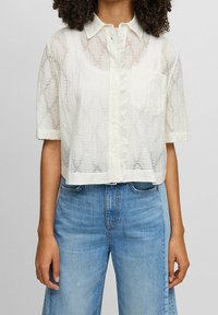 Vit kortärmad skjorta med krage, i randigt genomskinligt tyg, med knappar fram och en liten ficka på vänster bröst.