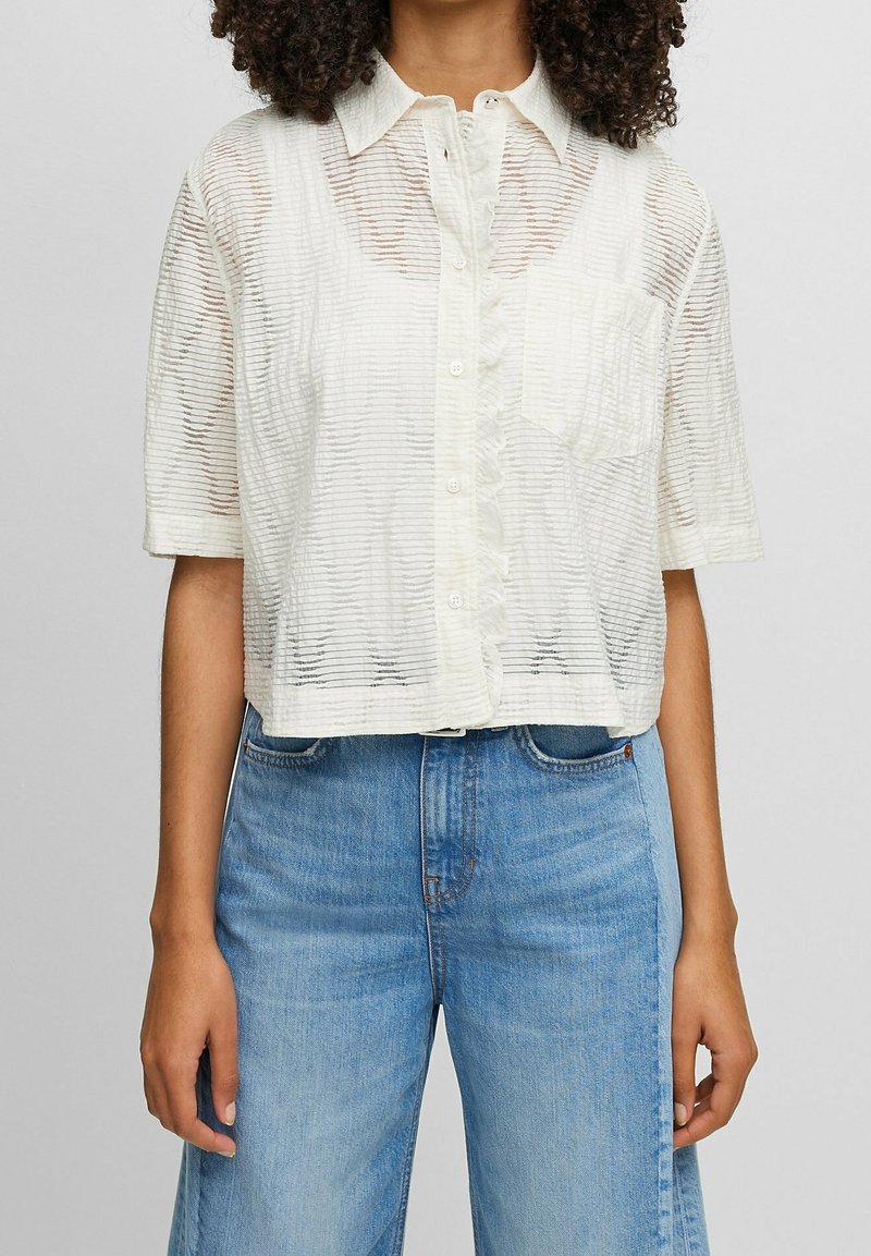 Vit kortärmad skjorta med krage, i randigt genomskinligt tyg, med knappar fram och en liten ficka på vänster bröst.