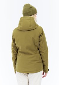 Osoba oblečená v olivovozelenom zateplenom plášti s kapucňou a zodpovedajúcou čiapkou, stojí so chrbtom otočeným k objektívu, na jednoduchom bielom pozadí.