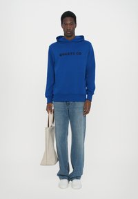 Felpa blu con logo "QUARTZ CO", tasca frontale, polsini a costine; indossata con jeans azzurri chiari e sneakers bianche, mentre tiene una borsa tote beige.