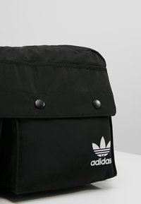 Svart Adidas tygväska med lock som är fäst med två svarta tryckknappar och vit Adidas-logotyp på framsidan.