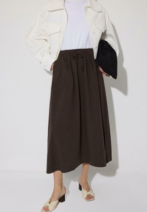 Osoba oblečená v bielom krátkom sakom, bielom topánke, tmavohnedej midi sukni, krémových sandáloch na opätku, držiaca čiernu listovú kabelku.