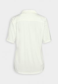 Chemise blanche à manches courtes avec un col, vue de dos sur un fond clair uni.