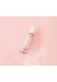 Rosa cylindrisk flaska med vit pump-lock. Etiketten har svart text. Slät yta, genomskinlig bas som visar vätskan inuti. Ställt mot en rosa bakgrund.