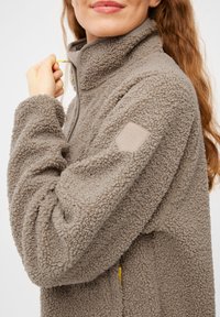 Fleece-Jacke in Taupe mit hohem Kragen, strukturiertem Stoff und einem Lederpatch am Ärmel. Der Reißverschluss-Zipper ist gelb.