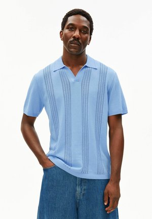 Mann trägt ein hellblaues, strukturiertes, kurzärmeliges Strickhemd mit Kragen und blaue Jeanshose, steht mit einer Hand in der Tasche.