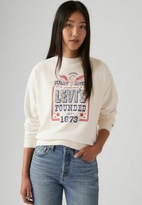 Felpa crema con stampa grafica che presenta il testo "Levi's Fondata nel 1873", abbinata a jeans in denim blu chiaro. Tessuto morbido, vestibilità rilassata.