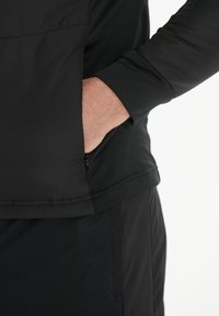 Sort, langærmet pullover med en glat tekstur. Har en frontlomme og en lynlåsdetaile i siden, parret med sorte bukser.