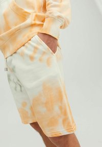 Szorty tie-dye z bawełny w jasnym kremowym kolorze z pomarańczowymi i zielonymi wirami, z boczną kieszenią oraz elastycznym pasem ze sznurkiem.