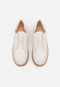 Chaussures en toile beige clair avec des lacets blancs, une finition en cuir texturé et une semelle en caoutchouc marron. Présentent des coutures minimales et un design épuré.