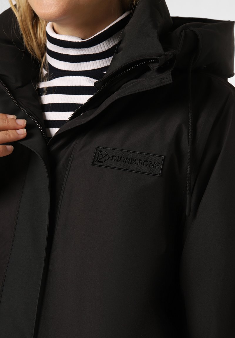 Schwarze wasserdichte Jacke mit Kapuze und Reißverschluss, aus strukturiertem Stoff gefertigt und mit einem schwarzen gestickten Logo auf der Brust.