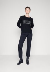 Maglione nero con dettaglio grafico testurizzato al centro, abbinato a pantaloni neri sartoriali e stivali neri con tacco. Finitura in tessuto liscia.