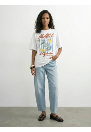 Kvinde iført hvid grafisk t-shirt med skaldyr og citron design, lyseblå jeans, brune loafers og armbånd, stående foran en ensfarvet baggrund.